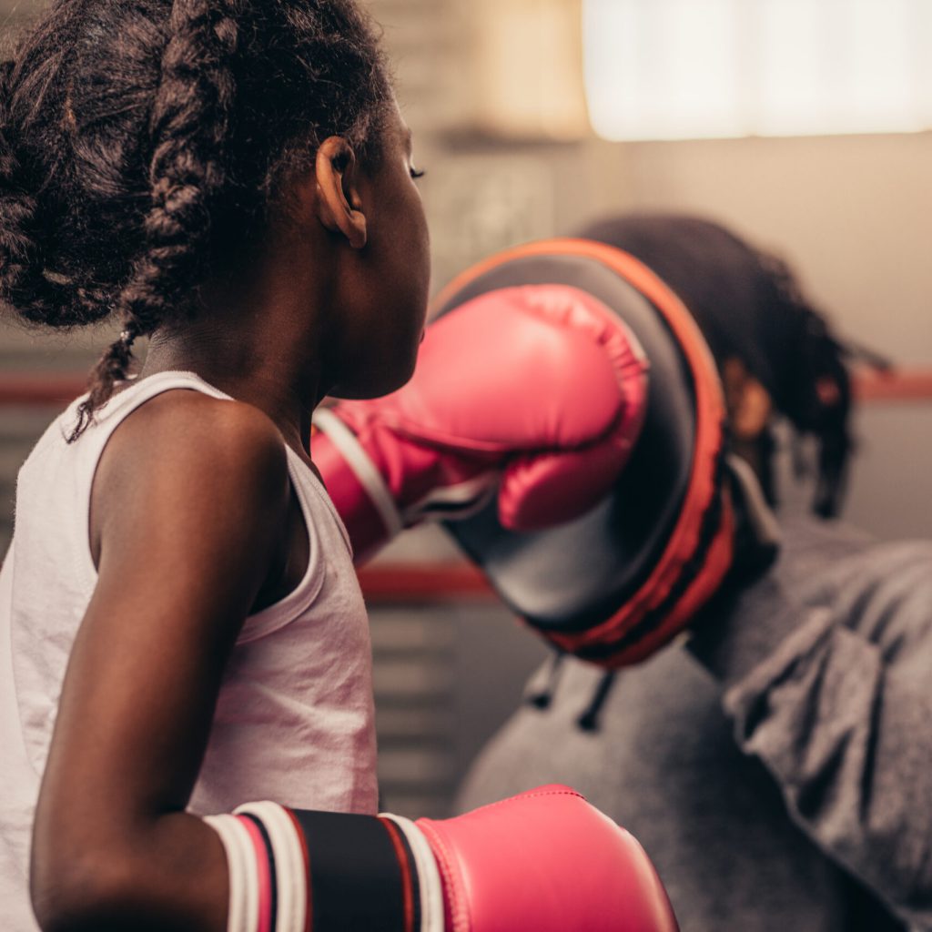 Girl wearing boxing gloves straining with her coach. Boxing kid practicing punches on a punching pad with her coach.